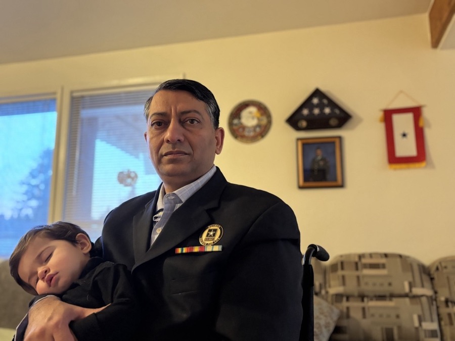 Zahid Chaudhry, seated, holding his sleeping child, in front of his military memorial wall at home